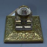 A cut glass inkwell (a/f), on a square polished brass stand decorated with scrolling foliage and