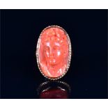 A yellow metal and coral cameo ring set with an oval coral plaque carved in the form of a lady's