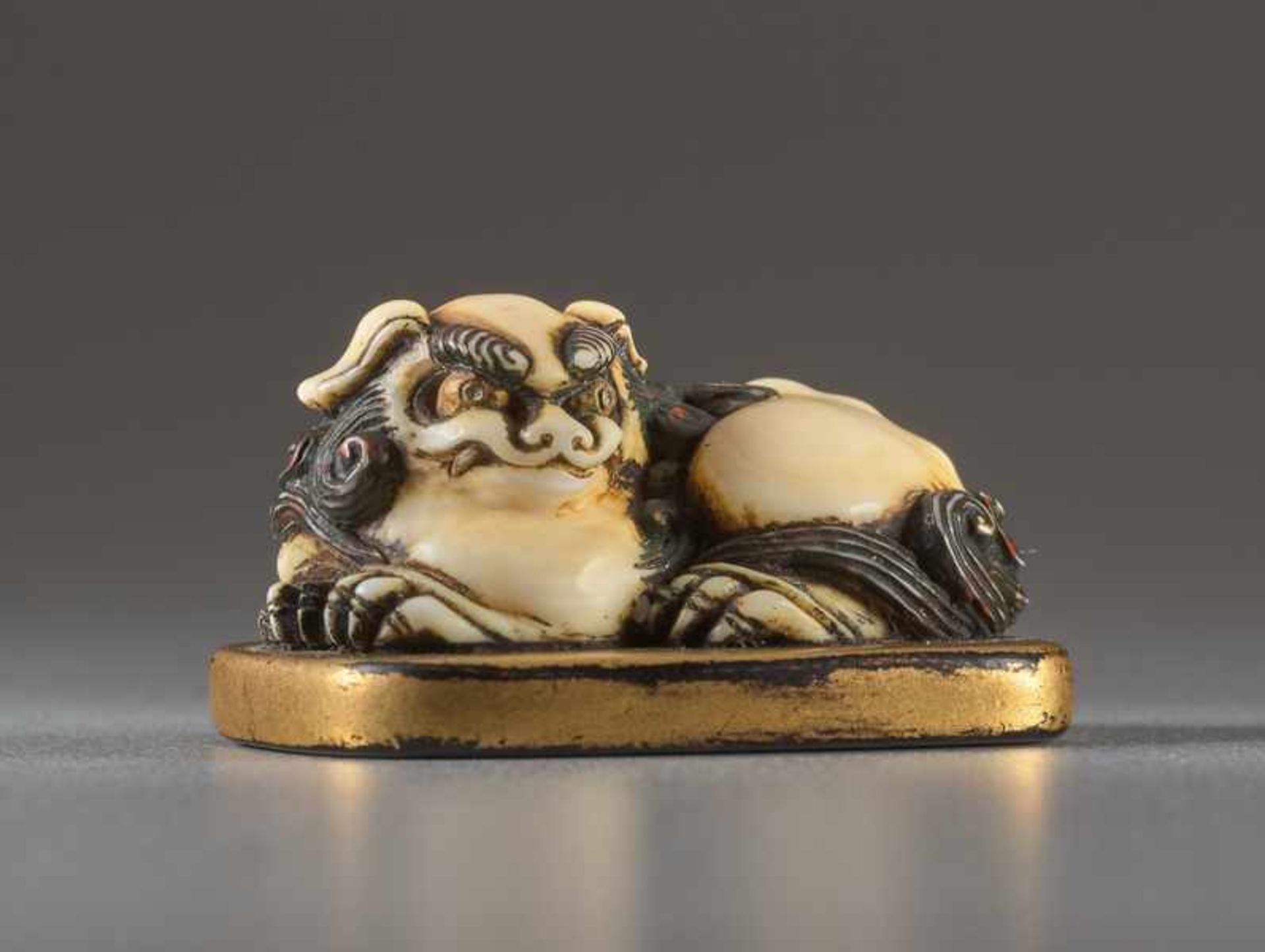 AN IVORY AND LACQUER NETSUKE OF A RECUMBENT SHISHI ON MAT Ivory netsuke with gold lacquer. Japan,