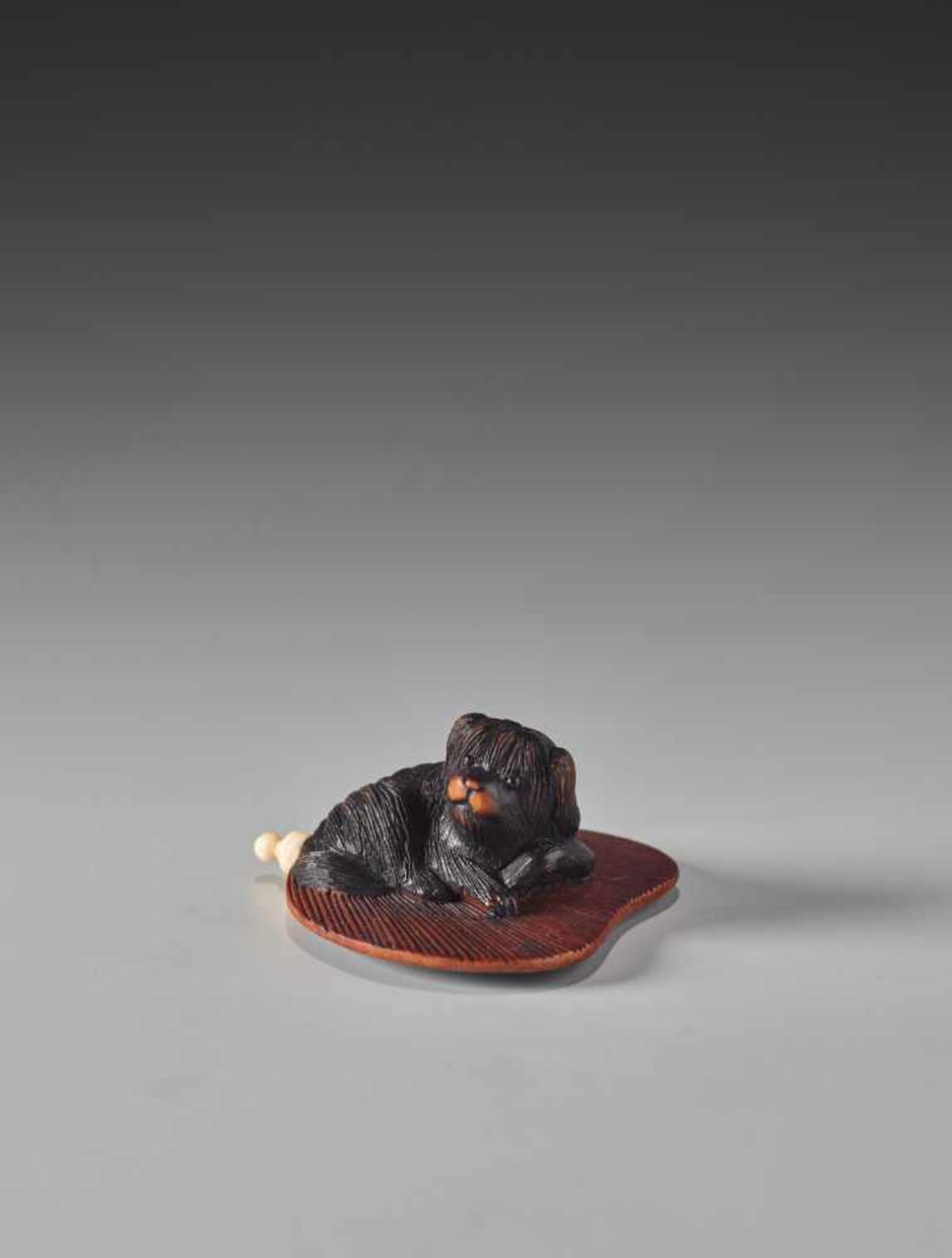 A FINE WOOD NETSUKE OF A PEKINGESE DOG ON A FAN WITH INSCRIBED LACQUER POEM BY MASATOMOWood