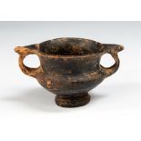 A black glazed pottery kantharos wine cup with a wide body and two ring handles surmounted by