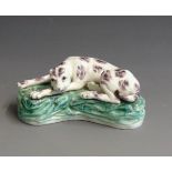 A pearlware model of a hound laying on a grassy mound, possibly an ink well or a candle holder,