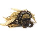 An Asmat tribal skull, with tusk front, beaded head and feathered top, the main body 17cm high.