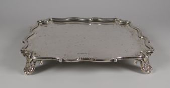 A SQUARE SILVER SALVER having a shaped scrolling border, raised over acanthus feet, Sheffield