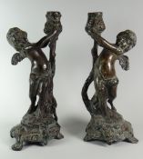 A PAIR OF BRONZE CHERUBIC CANDLEHOLDERS on naturalistic footed bases, 29cms high