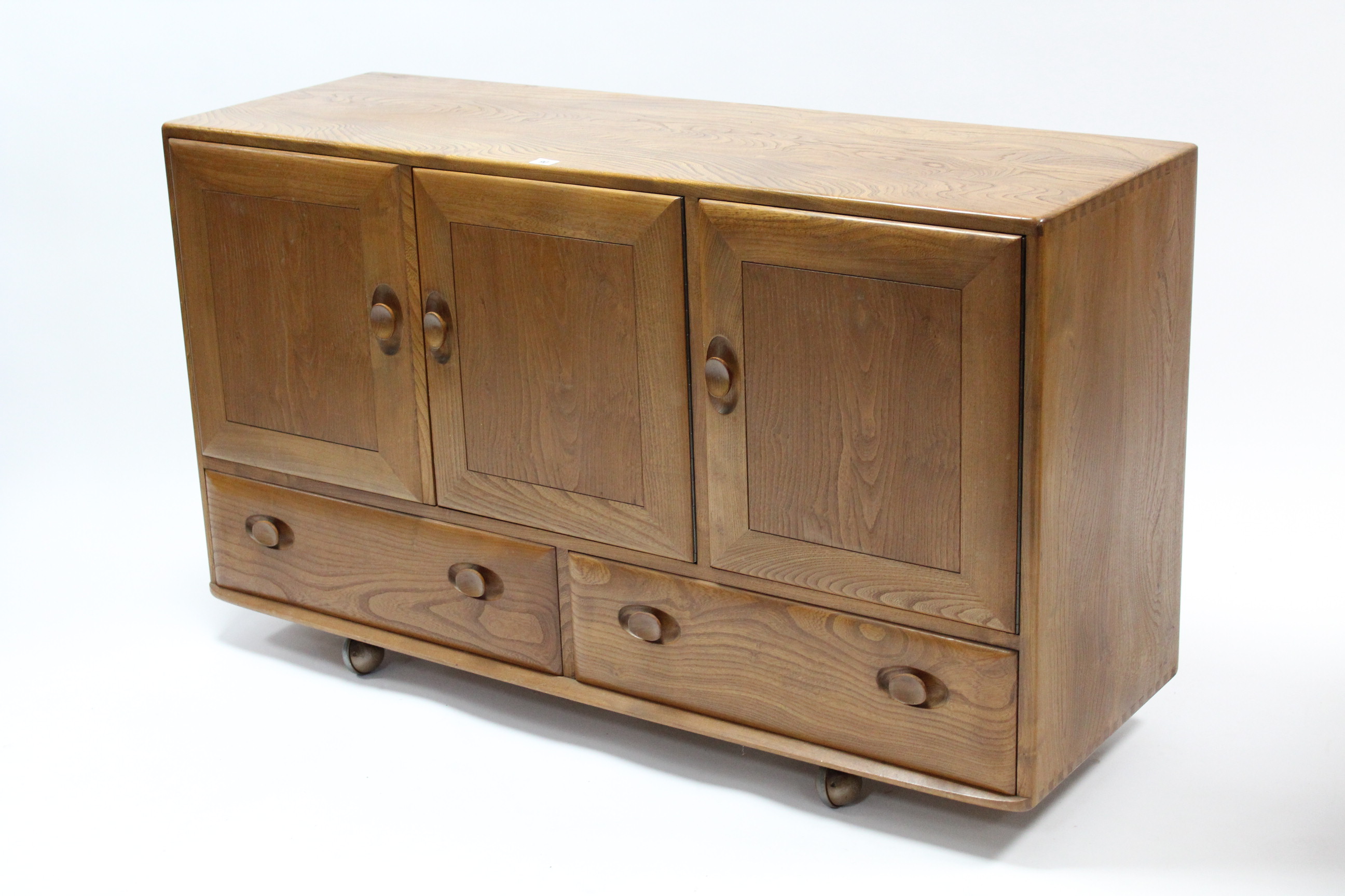 AN ERCOL LIGHT ELM SIDEBOARD enclosed by three panel doors above two short drawers, & on Shepherd’