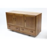 AN ERCOL LIGHT ELM SIDEBOARD enclosed by three panel doors above two short drawers, & on Shepherd’