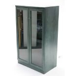 A green-finish wooden side cabinet enclosed by pair of glazed doors, & on plinth base, 30” wide x
