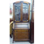 A 1930s oak bureau bookcase, the arched upper part enclosed by a pair of lead glazed panel doors,