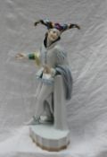 A Karl Ens porcelain figure of a Jester, with multicoloured tricorn hat leaning against a pillar,