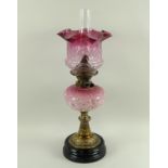 An oil lamp with a cranberry glass shade, a pink ceramic well, brass stem and turned slate base,