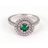 A platinum, emerald and diamond ring, the round emerald within a double diamond halo,