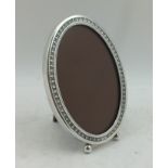 AN OVAL SILVER PHOTOGRAPH FRAME, decorative edge, two ball feet to base, polished wood easel back,