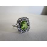 A white coloured metal ring set with a peridot within a border of fourteen diamonds, 15mm x 13mm,