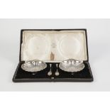 CASED PAIR OF GEORGE V SHELL SHAPED SILVER BUTTER DISHES AND KNIVES, of typical form with ball feet,