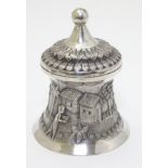 A white metal pot and cover with decoration to sides of figures and village buildings.