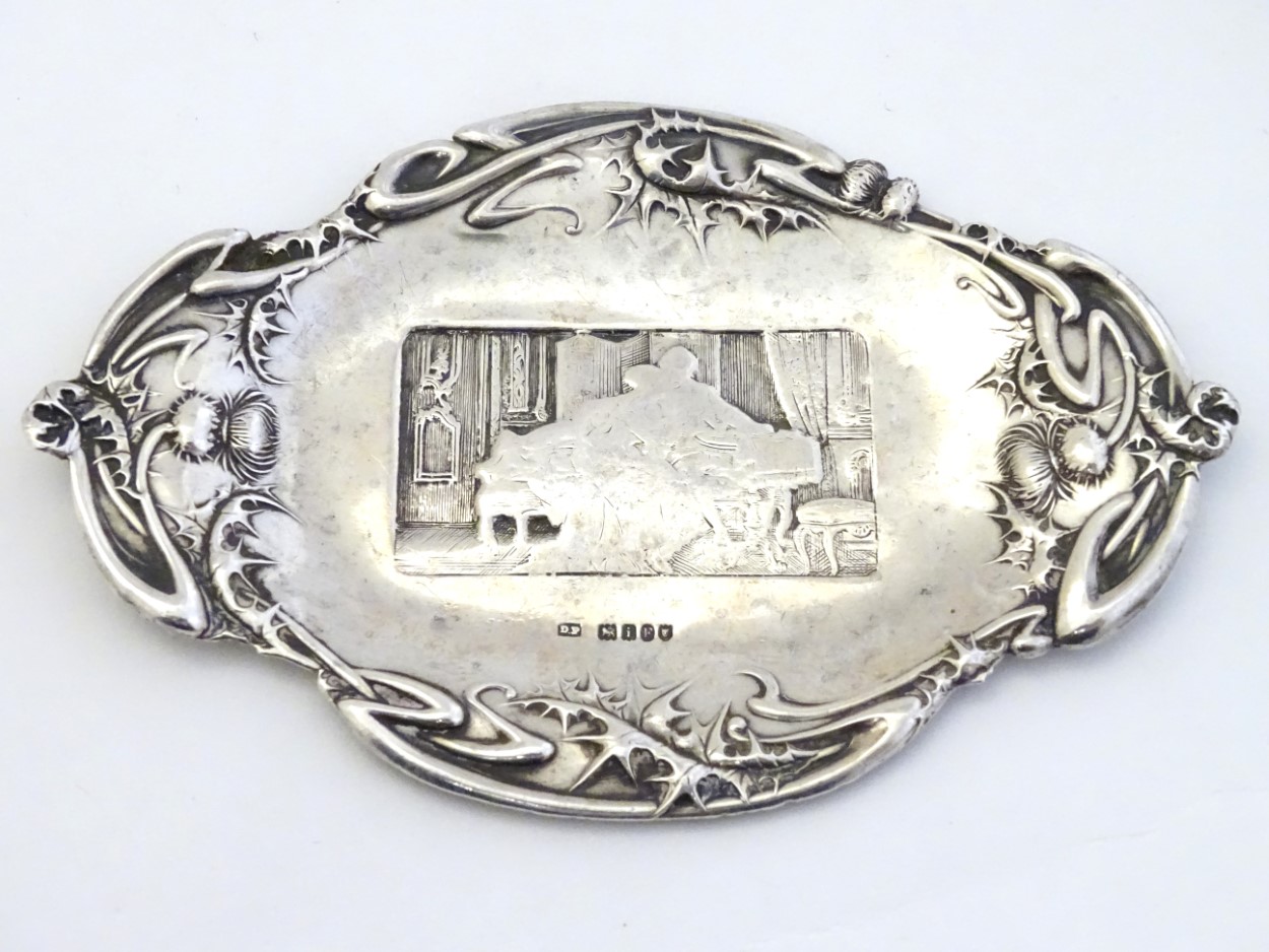 A silver pin dish with thistle decoration to border and interior scene to centre.