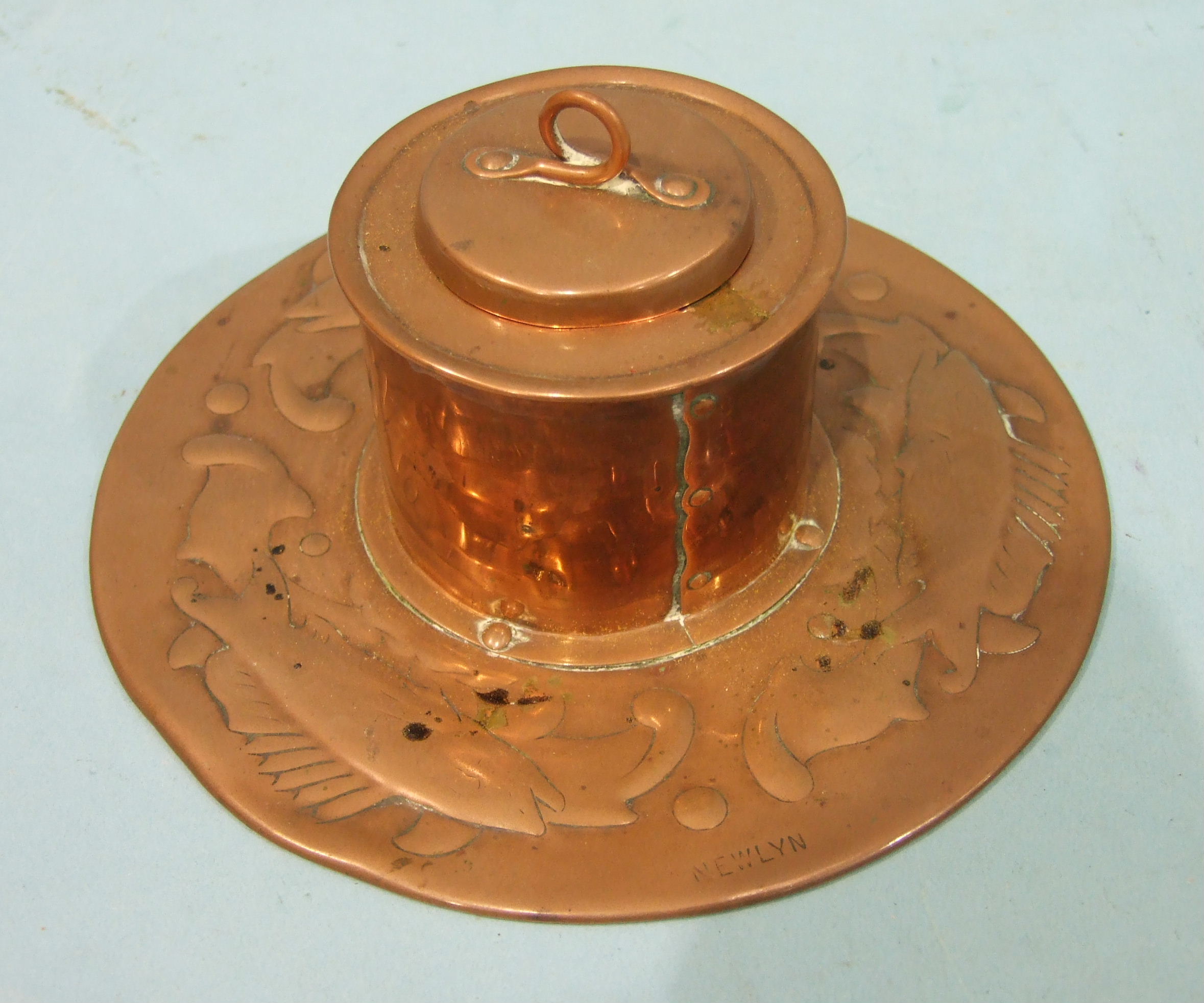 A Newlyn copper inkwell of cylindrical form, with lid, on a circular base with embossed fish