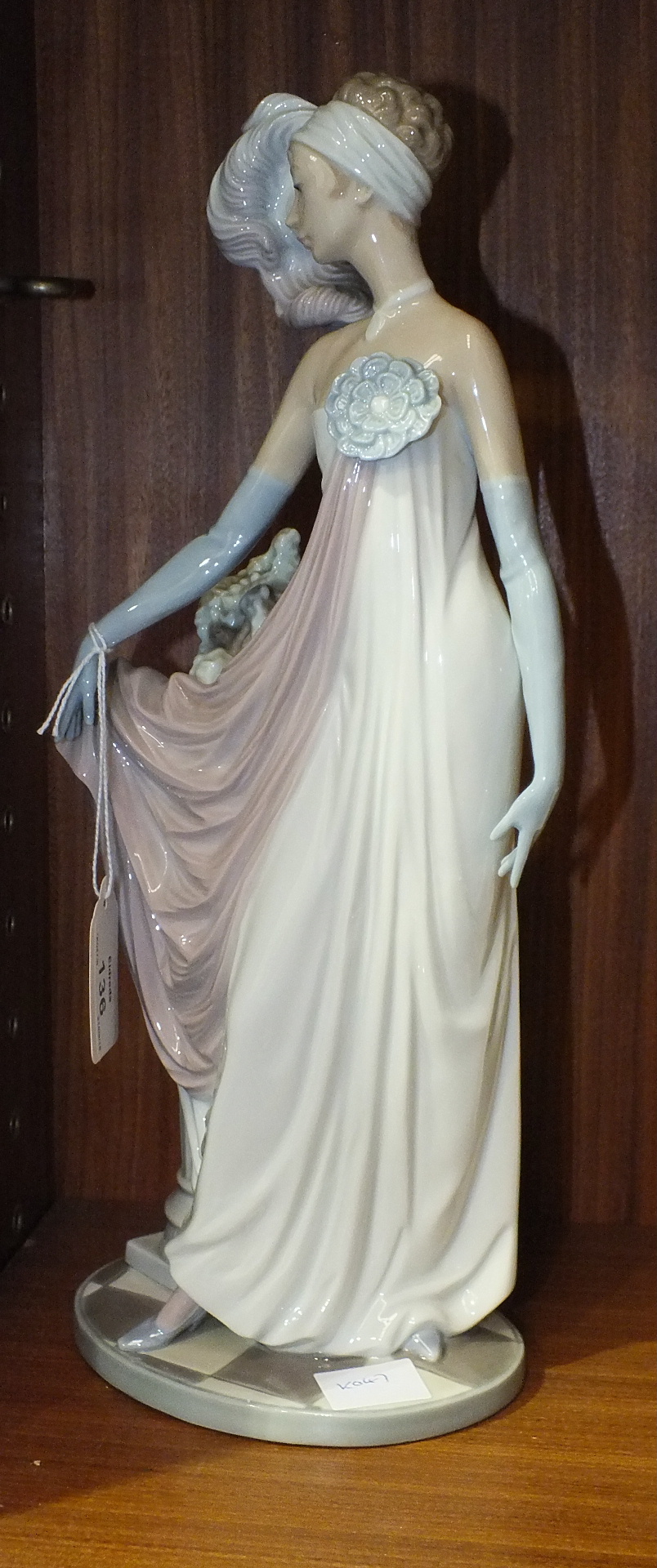 A Lladro figure of a young woman with plumed head dress and evening gown standing by a pedestal of