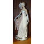 A Lladro figure of a young woman with plumed head dress and evening gown standing by a pedestal of