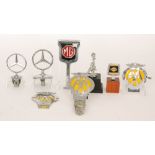 Two chromed Mercedes adjustable car badges, a MG variant, a Nissan badge on a wooden plinth,