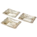 A composed set of three hallmarked silver shaped rectangular dishes with acanthus scroll borders,