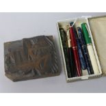 A woodblock carved with a printers press and various vintage fountain pens