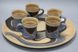 An Usch Spettigue pottery wall plaque and a set of five mugs, all of similar linear design in