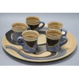 An Usch Spettigue pottery wall plaque and a set of five mugs, all of similar linear design in