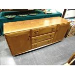 A LIGHT ERCOL SIDEBOARD fitted with three drawers flanked by cupboards, supported on four casters,