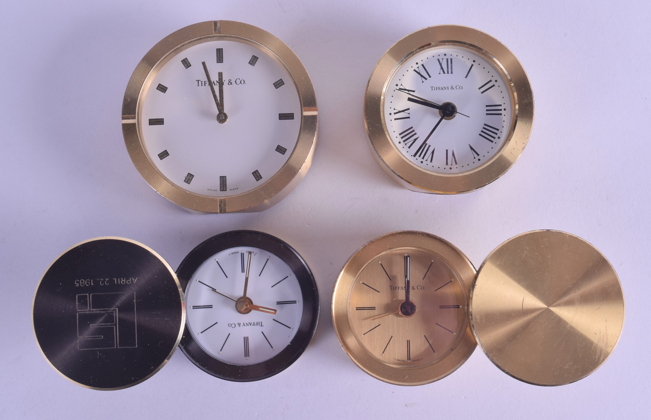 FOUR TIFFANY & CO BRASS TRAVELLING CLOCKS in various forms and sizes. Largest 7 cm diameter. (4)