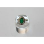 An emerald and diamond cluster ring the oval cut stone claw set in a border of twelve brilliant