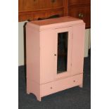 A pink painted wooden doll's wardrobe with central mirrored panel door and drawer below, 62.5cm by