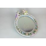 An oval porcelain wall mirror decorated with two cherubs and flower encrusted frame, bevelled mirror