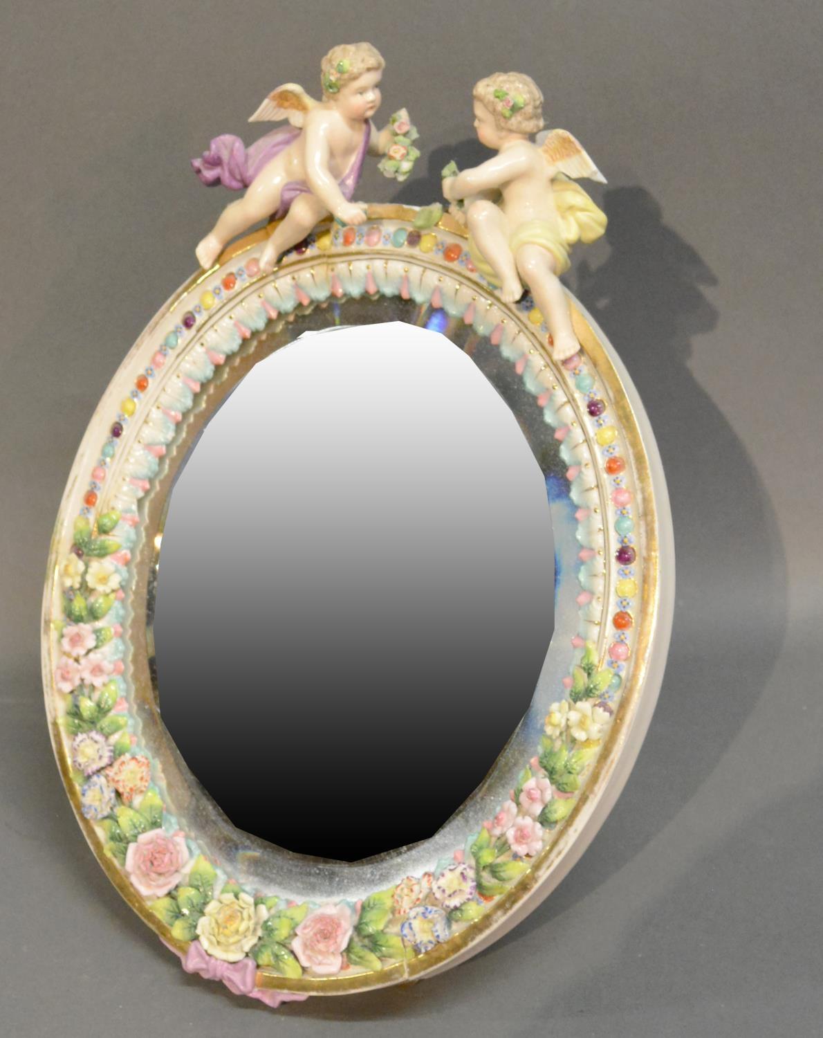 A German Porcelain Oval Table Mirror Surmounted With Cupid and foliate encrusted decoration, 36cm by