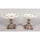 A PAIR OF SILVER AND OPAQUE GLASS COMPORTS, the shaped glass tops with gilded decoration on a rococo