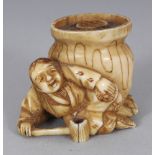 A JAPANESE MEIJI PERIOD STAINED IVORY NETSUKE OF A MAN RECLINING BEFORE A CAULDRON, his ladle