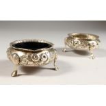 A PAIR OF VICTORIAN SALTS, repousse with flowers, on three curving legs, one liner. 2.75ins