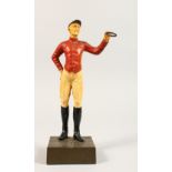 A PAINTED BRONZE STANDING FIGURE OF A JOCKEY, in a red jacket, on a square base. 10.5ins high.