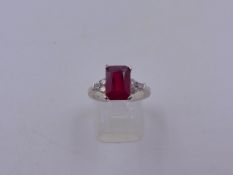 A 750 STAMPED WHITE METAL RUBY AND DIAMOND RING. THE CENTRAL RUBY IS AN EMERALD CUT IN AN ELEVATED