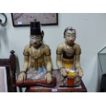 A PAIR OF MALAYSIAN PAINTED WOOD FIGURES OF A SEATED MAN AND HIS WIFE WEARING BLACK FEZ TYPE HATS,