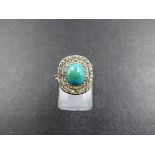 A WHITE METAL TURQUOISE AND DIAMOND RING. THE CENTRAL CABOCHON TURQUOISE IS SURROUNDED BY A DOUBLE