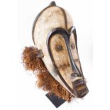 FANG FACE MASK, carved and whitened wood, with raffia embellishment, 52cm H, plus a display stand.