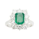 An emerald and diamond cluster ringclaw set with a trap cut emerald, in a single border of round