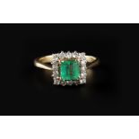 AN EMERALD AND DIAMOND CLUSTER RING, the rectangular step-cut emerald claw set within a border of