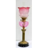 AN EDWARDIAN BRASS CORINTHIAN COLUMN OIL LAMP WITH EMBOSSED ROUND FOOT, CRANBERRY GLASS FOUNT AND