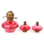 A CRANBERRY GLASS OIL LAMP FOUNT WITH BRASS BURNER AND TWO BRASS MOUNTED CRANBERRY PEG LAMPS, 19 AND