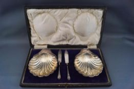 A pair of silver shell-shaped butter dishes each on three ball feet, with two small butter knives,