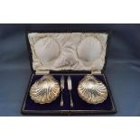 A pair of silver shell-shaped butter dishes each on three ball feet, with two small butter knives,