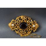 A yellow gold brooch set with a central oval garnet and two seed pearls. Ornate scroll design.